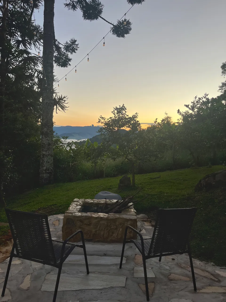 Duas cadeiras ao redor do fogo de chão, no gramado, com o sol se pondo atrás das árvores.
