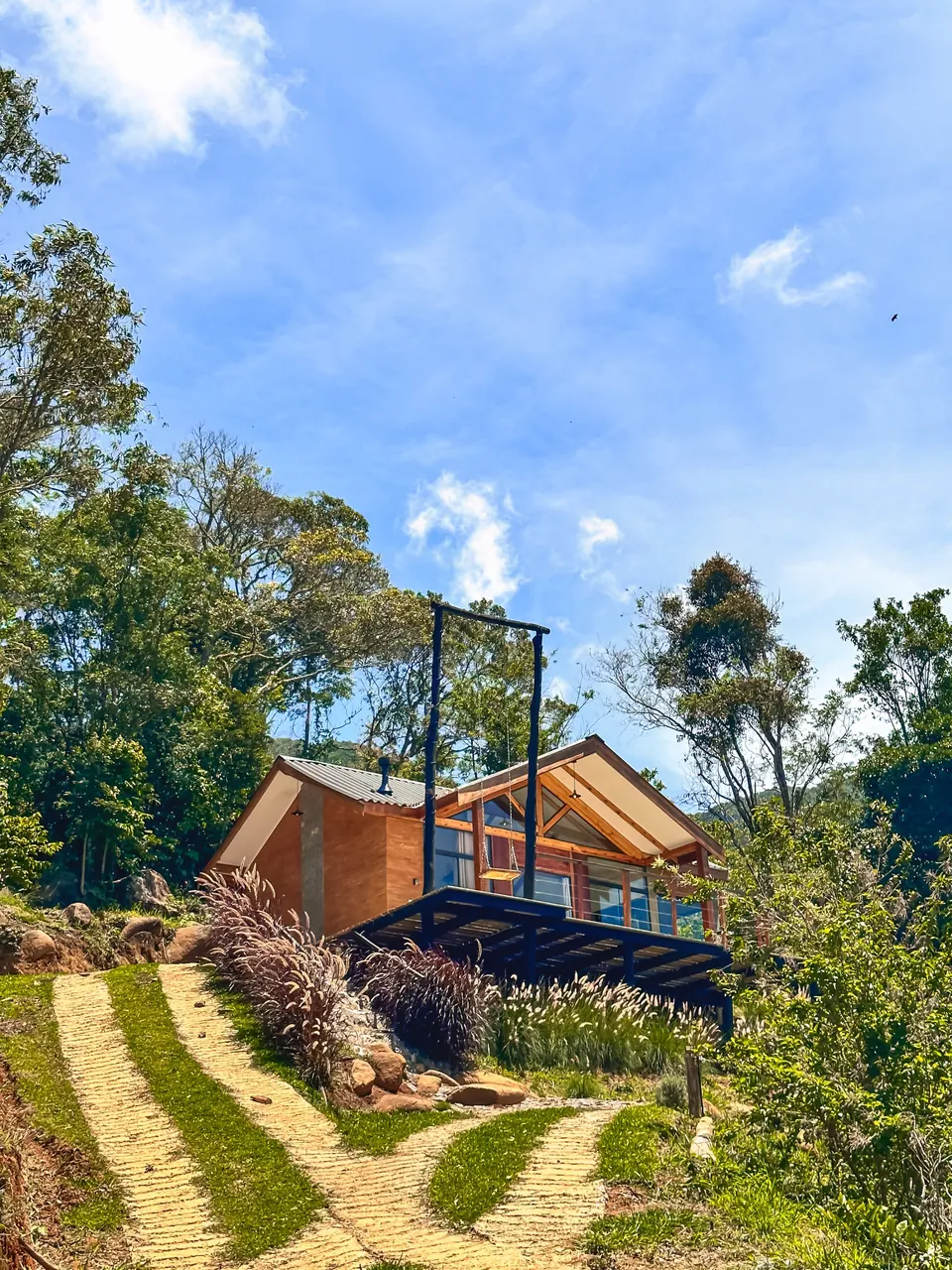 Belvedere de tijolo e vidro entre a mata, com um balanço ao lado e caminho de pedra na frente.