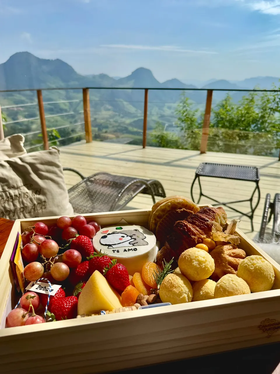 Cesta de café com frutas, pães de queijo, croissants e queijo, no deck com vista.