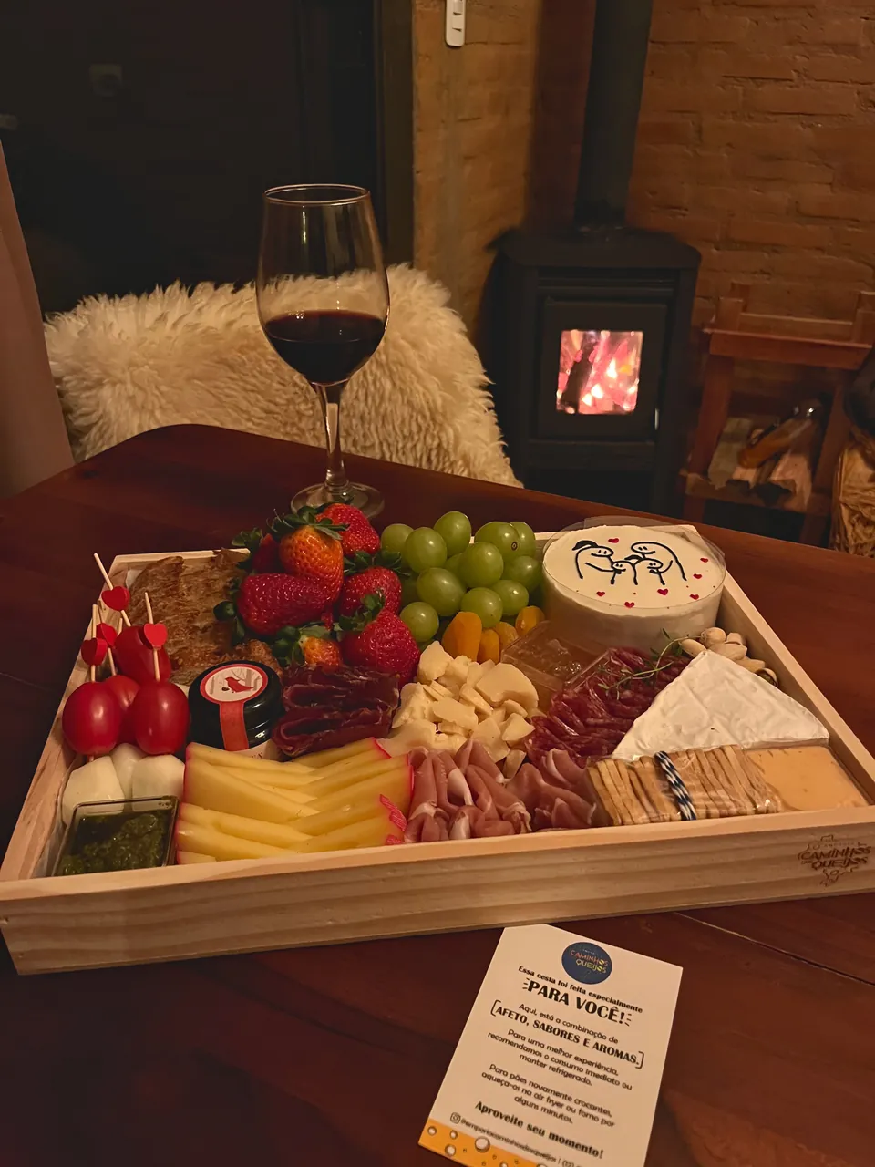 Tábua de frios e frutas com taça de vinho, perto de uma lareira acesa.