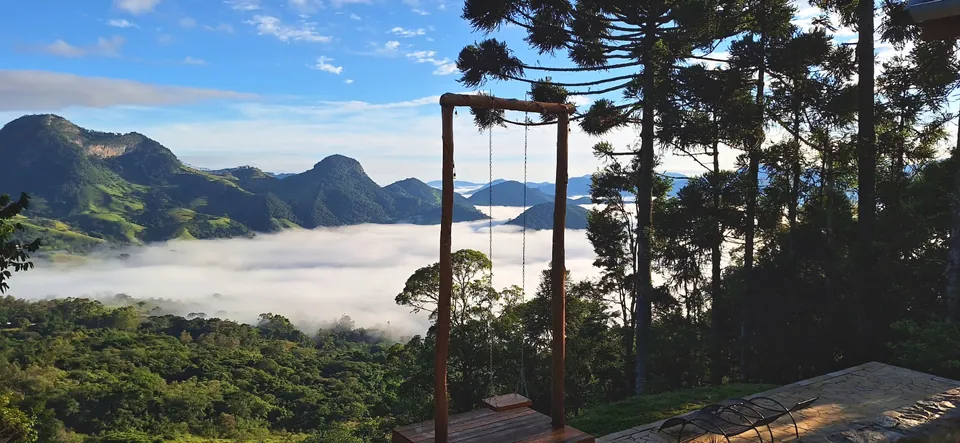 Balanço de madeira entre araucárias, com a névoa no vale e as montanhas ao fundo.