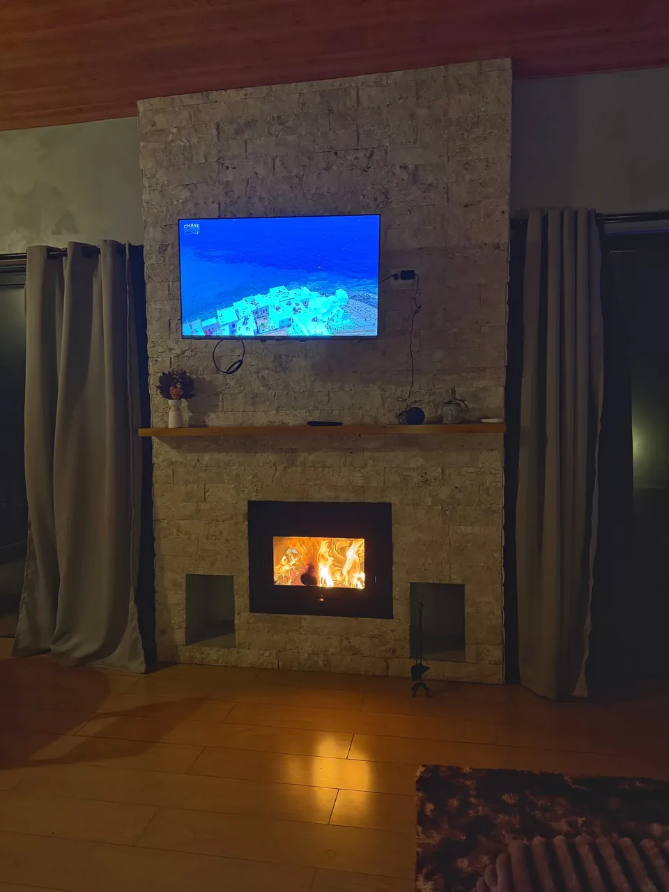 Lareira acesa na parede de pedra da Mantiqueira, com TV acima e cortinas dos lados.