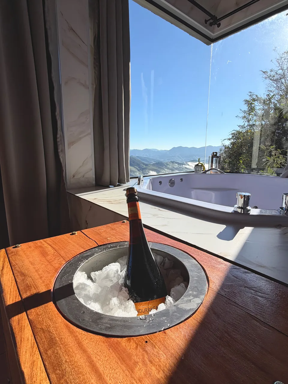 Garrafa no balde de gelo embutido na mesa de madeira, ao lado da banheira e da janela.